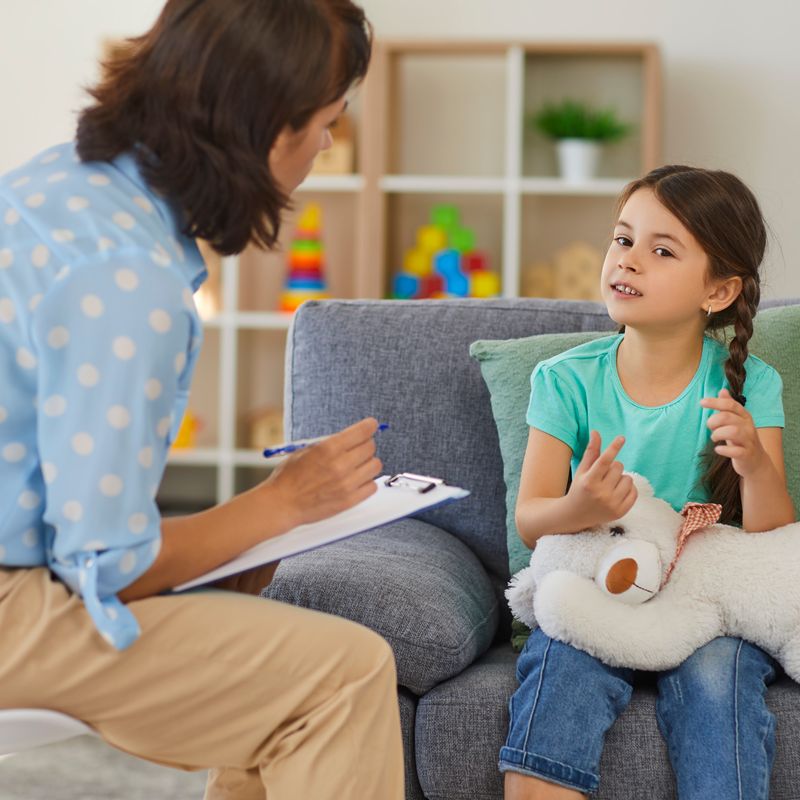 Supportive psychologist with clipboard listening to little child during therapy session. Preschool girl feeling at ease in therapist's office sharing her thoughts and concerns