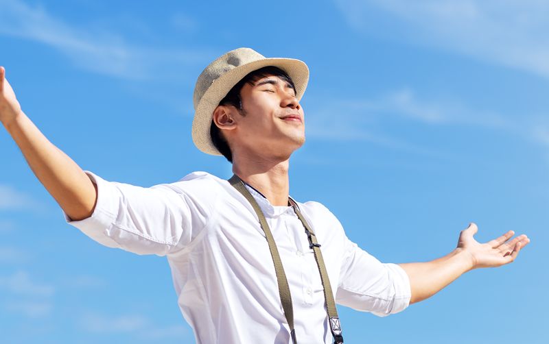 Candid of young attractive asian man close eyes, deep breath and wide open arms in blue sky view background feeling wind and sunshine. Happy asian hipster male in youth freedom culture travel concept.